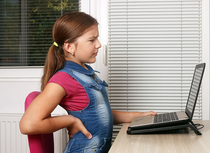 Ból pleców u dziecka - siedząca przy komputerze dziewczynka trzymająca się za bolące plecy